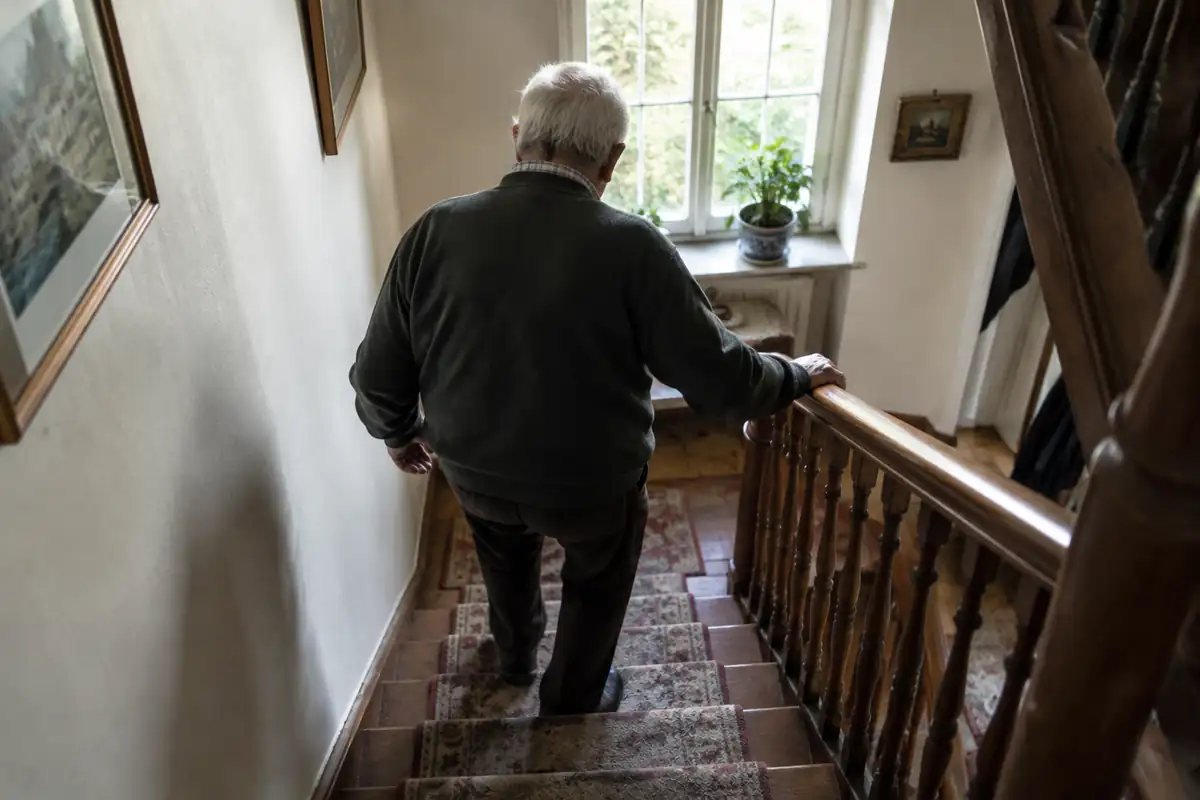 Homme âgé vu de dos qui descend un escalier de maison en tenant la rampe