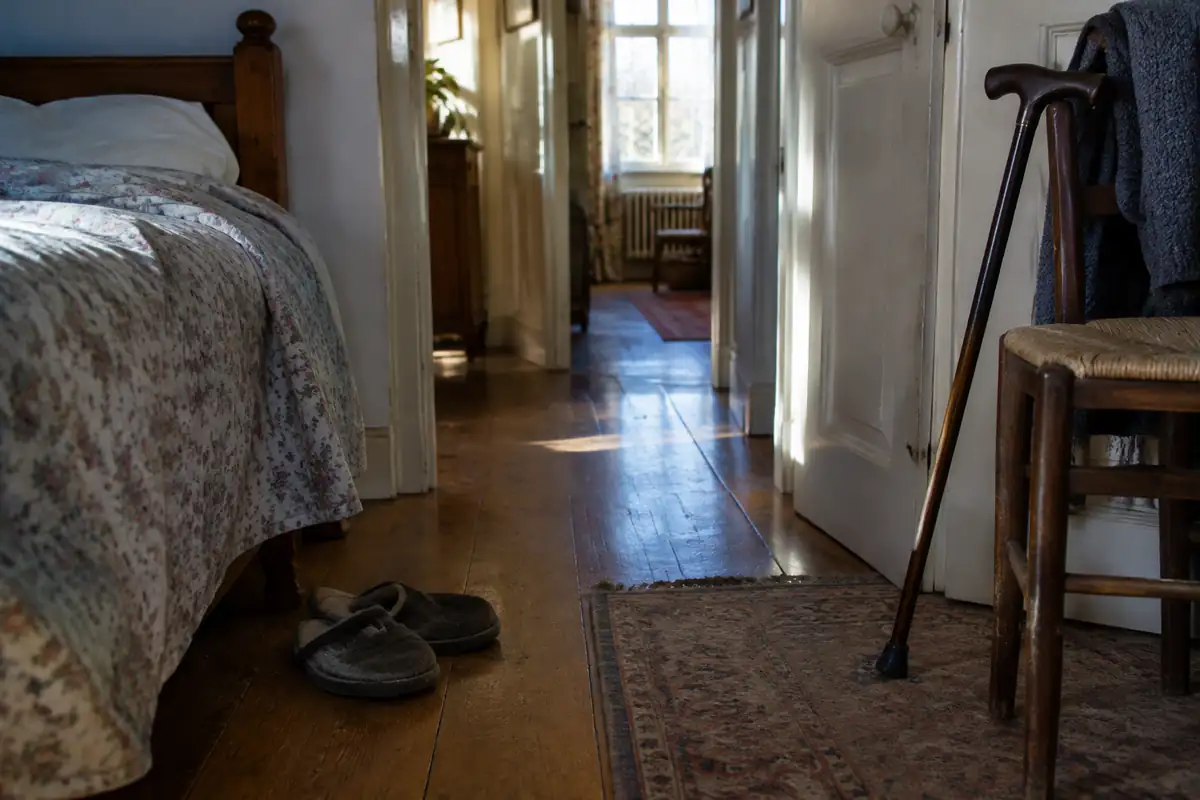 Pantoufles usées au bord d'un lit et canne appuyée contre une chaise dans une chambre au petit matin